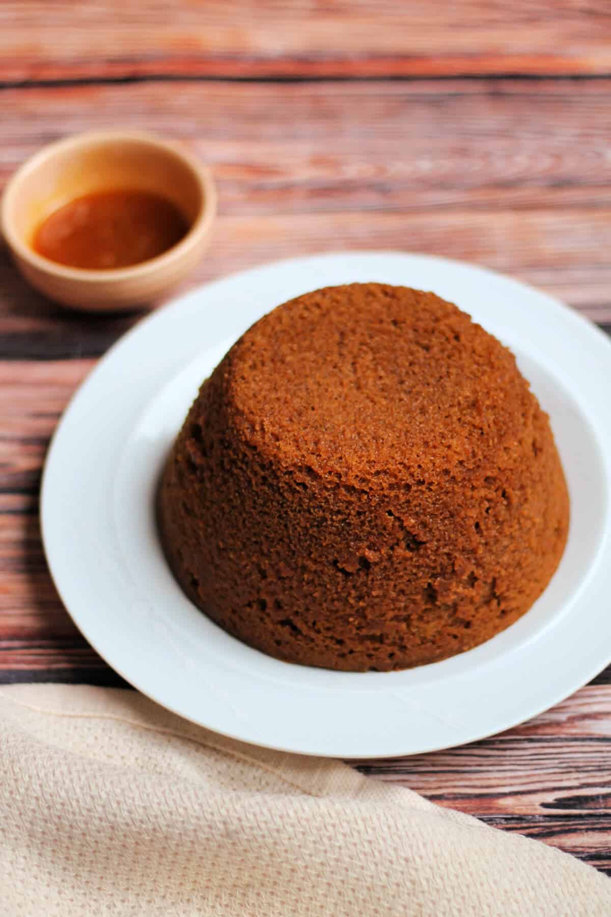 A whole gingerbread pudding on a white plate with a small dish of syrup behind.