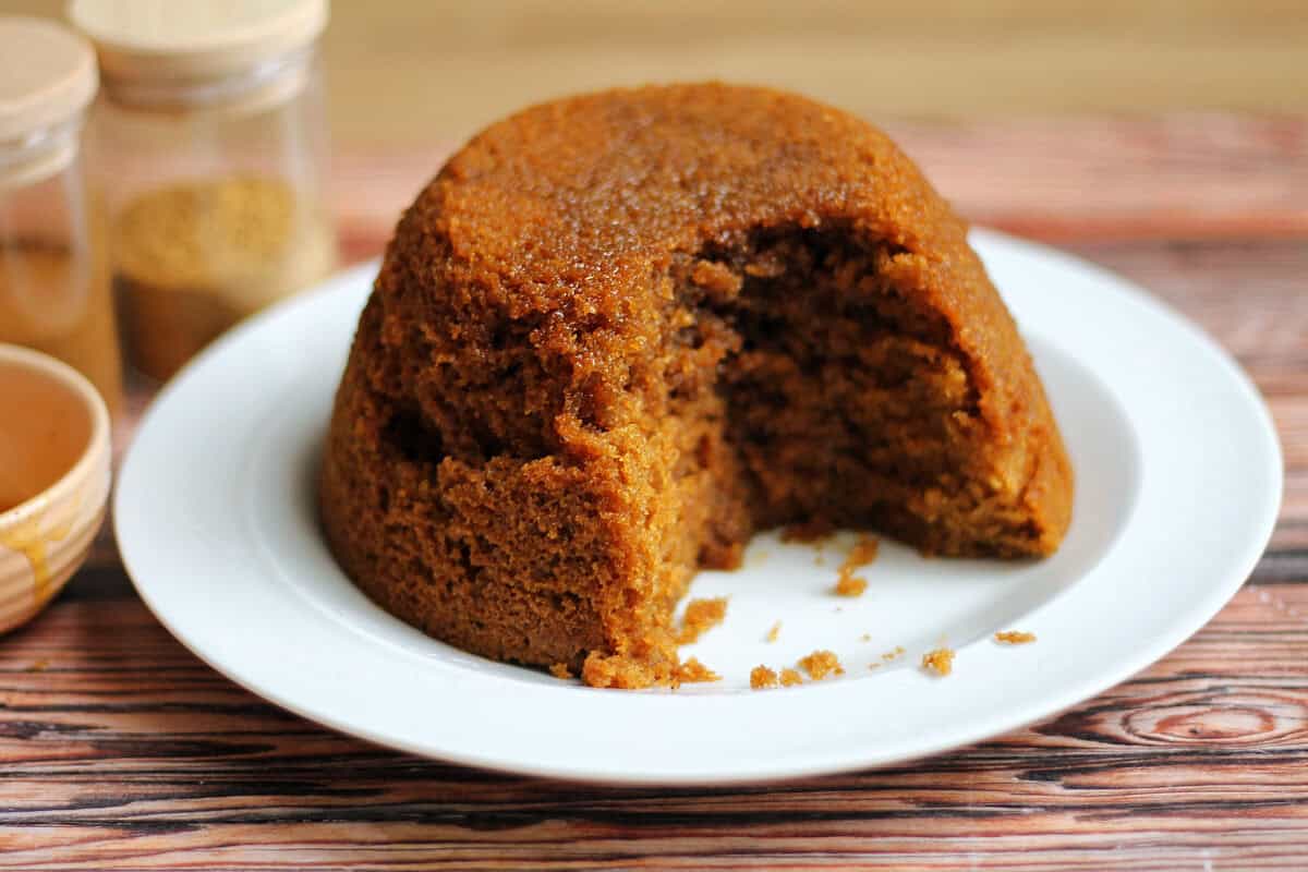 Gingerbread pudding on a white plate, with spice jars behind, a portion removed showing the soft centre.