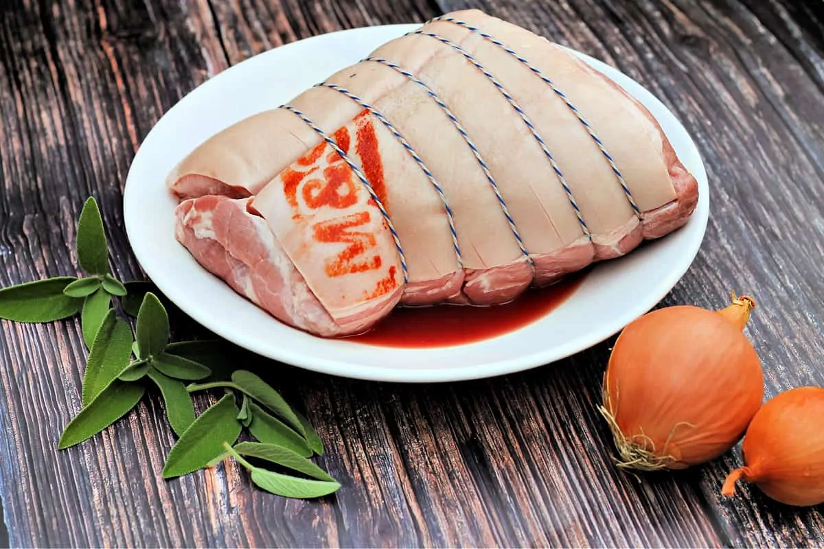 Joint of pork in a white bowl, with sage leaves and onions at the side on wooden surface.