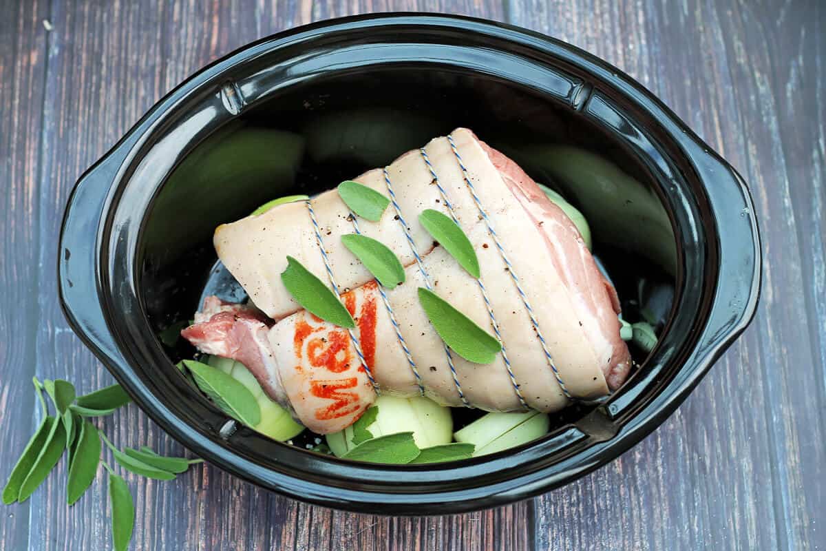 Joint of pork in slow cooker pot with salt, pepper and sage leaves on top and onions under it.