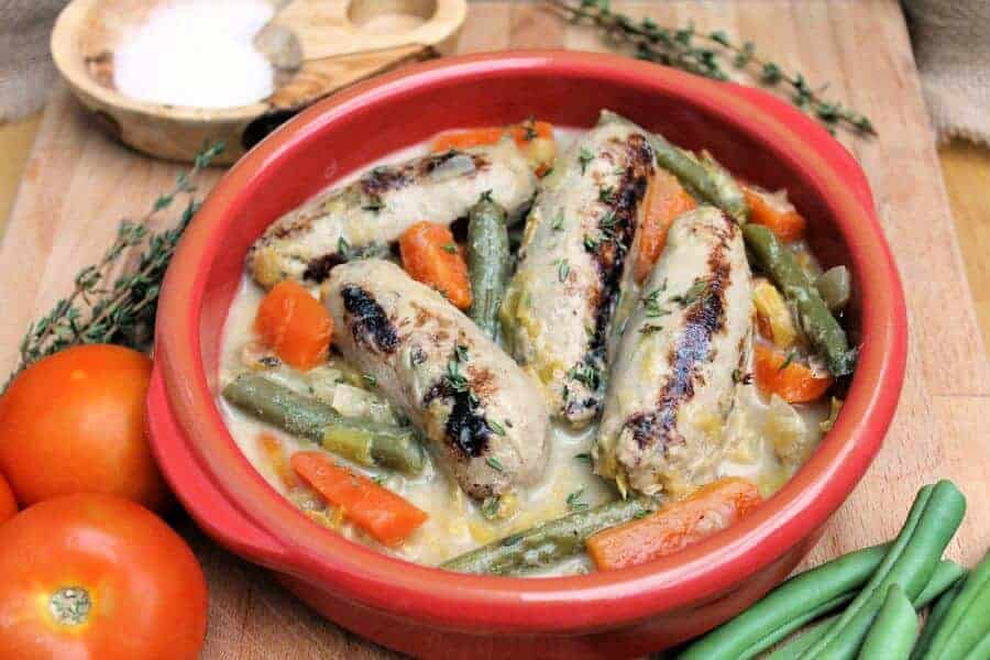 Red bowl with sausages and carrots, with tomatoes and green beans around it.