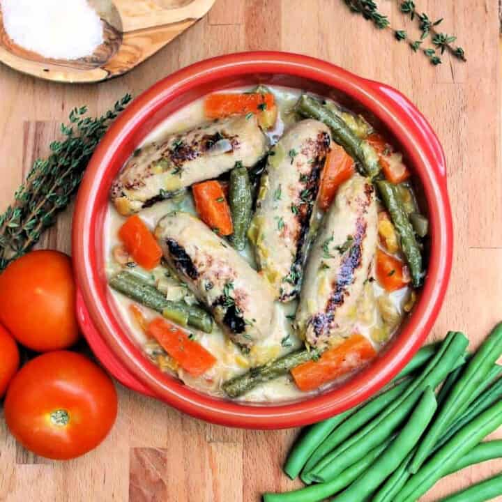 View from above of red bowl with sausages and carrots, with tomatoes, bowl of salt, herbs and green beans around it.