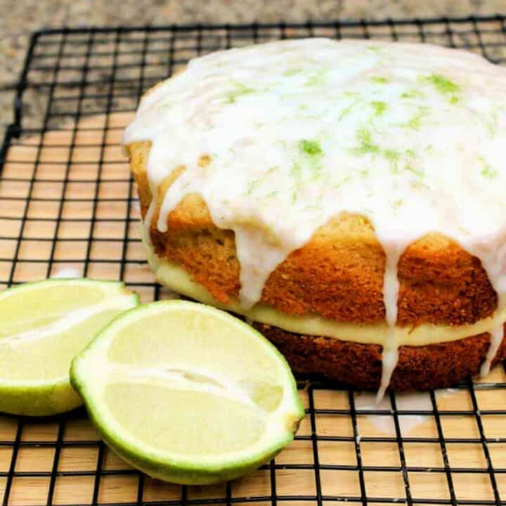Cake with drizzled white icing with limes beside it on a rack.