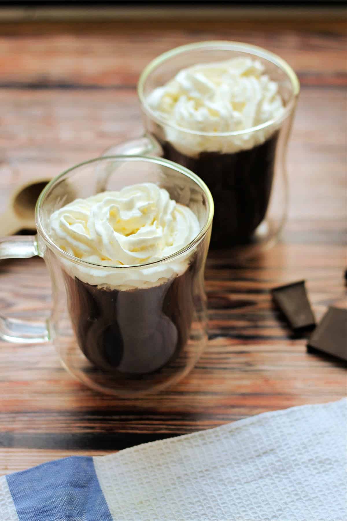 Two cups of hot chocolate with cream on top, with squares of chocolate beside them.
