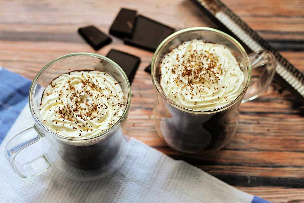 Two clear glass mugs of hot chocolate topped with whipped cream and chocolate shavings, with squares of chocolate and zester behind.