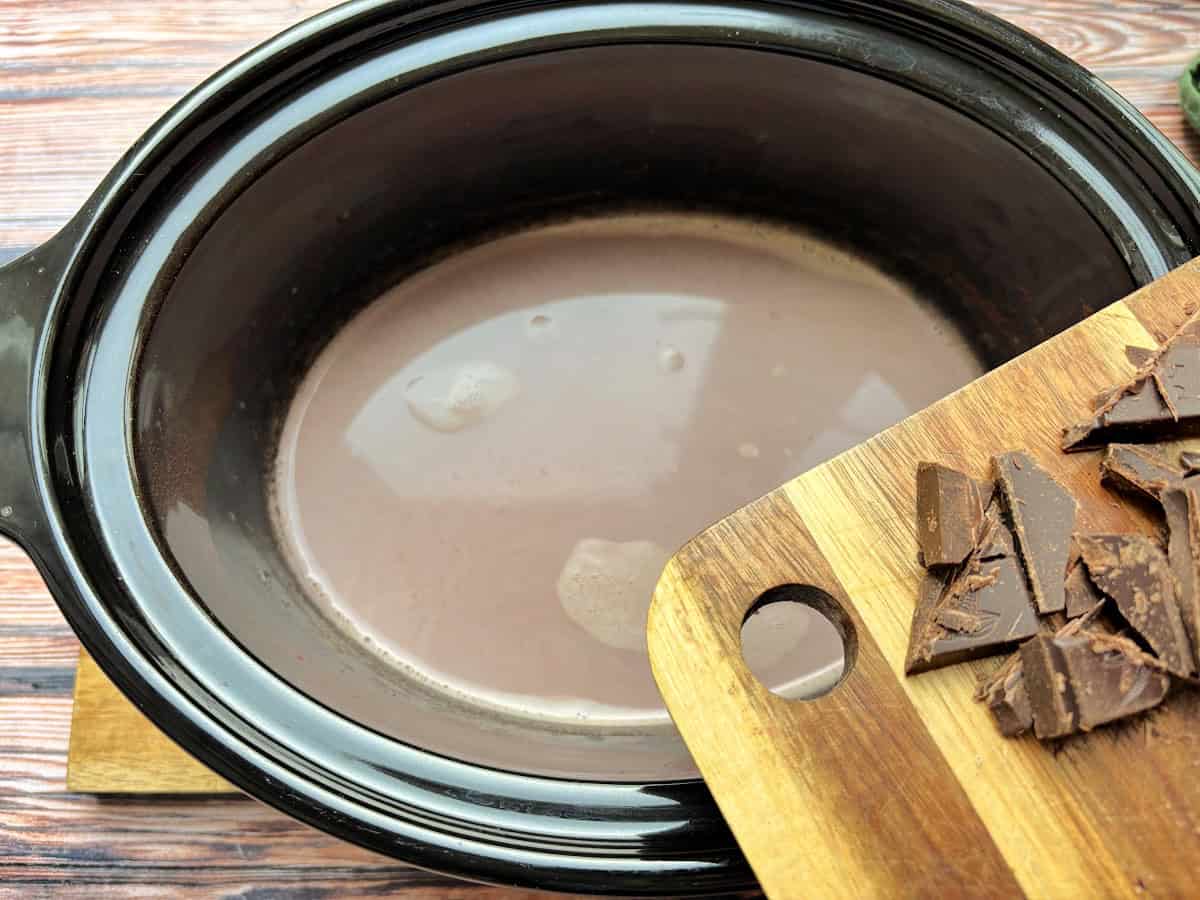 Squares of chocolate on a wooden board, about to be added to slow cooker pot with hot chocolate in it.