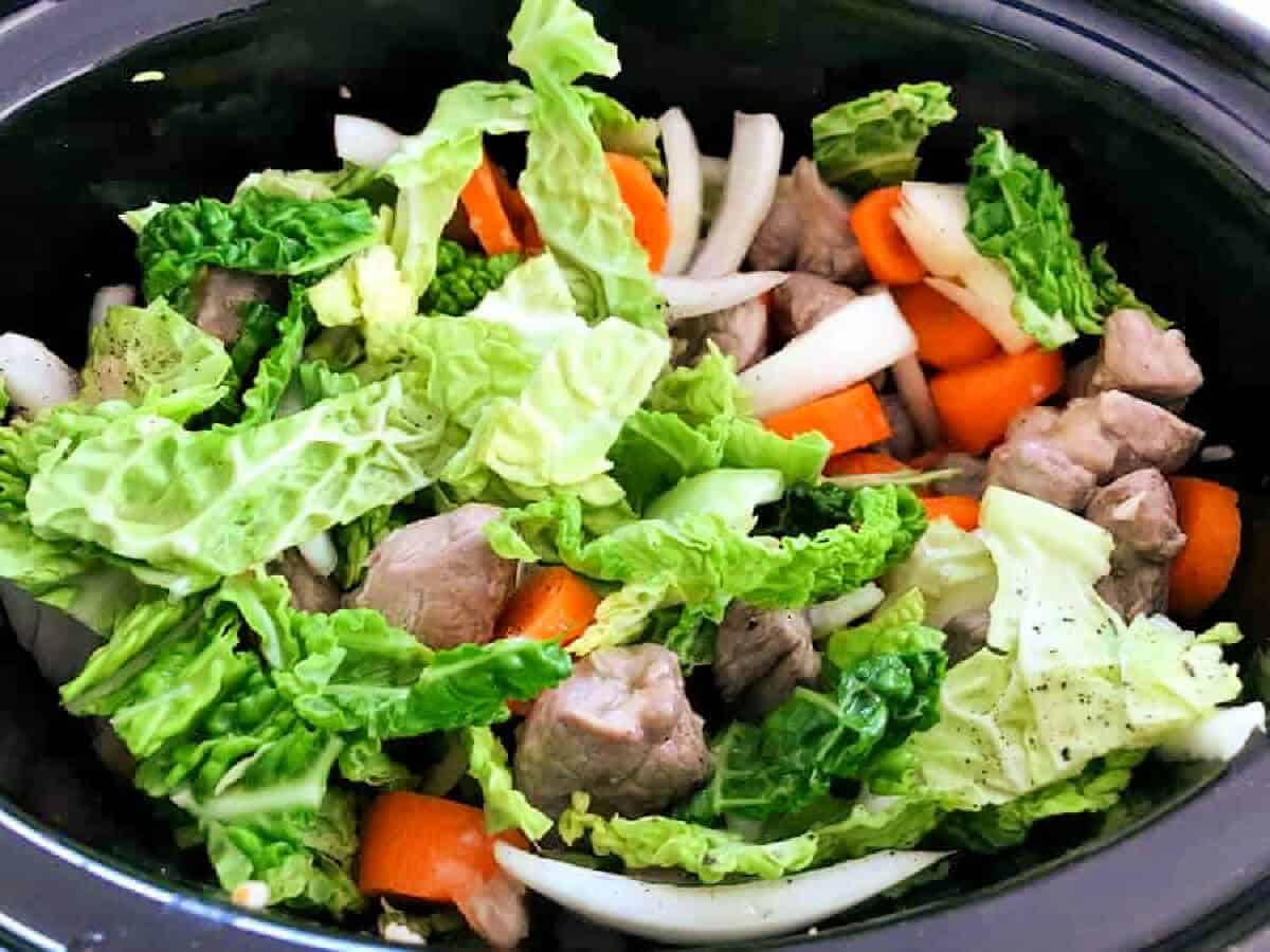 Slow cooker pot with cabbage, onion, carrot and lamb stew ingredients.