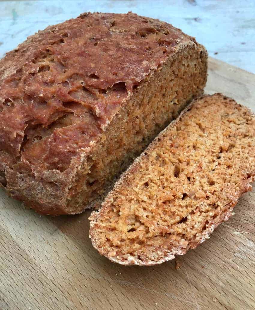 A loaf of bread on a wooden chopping board, with a slice cut out, showing the reddish bread inside.