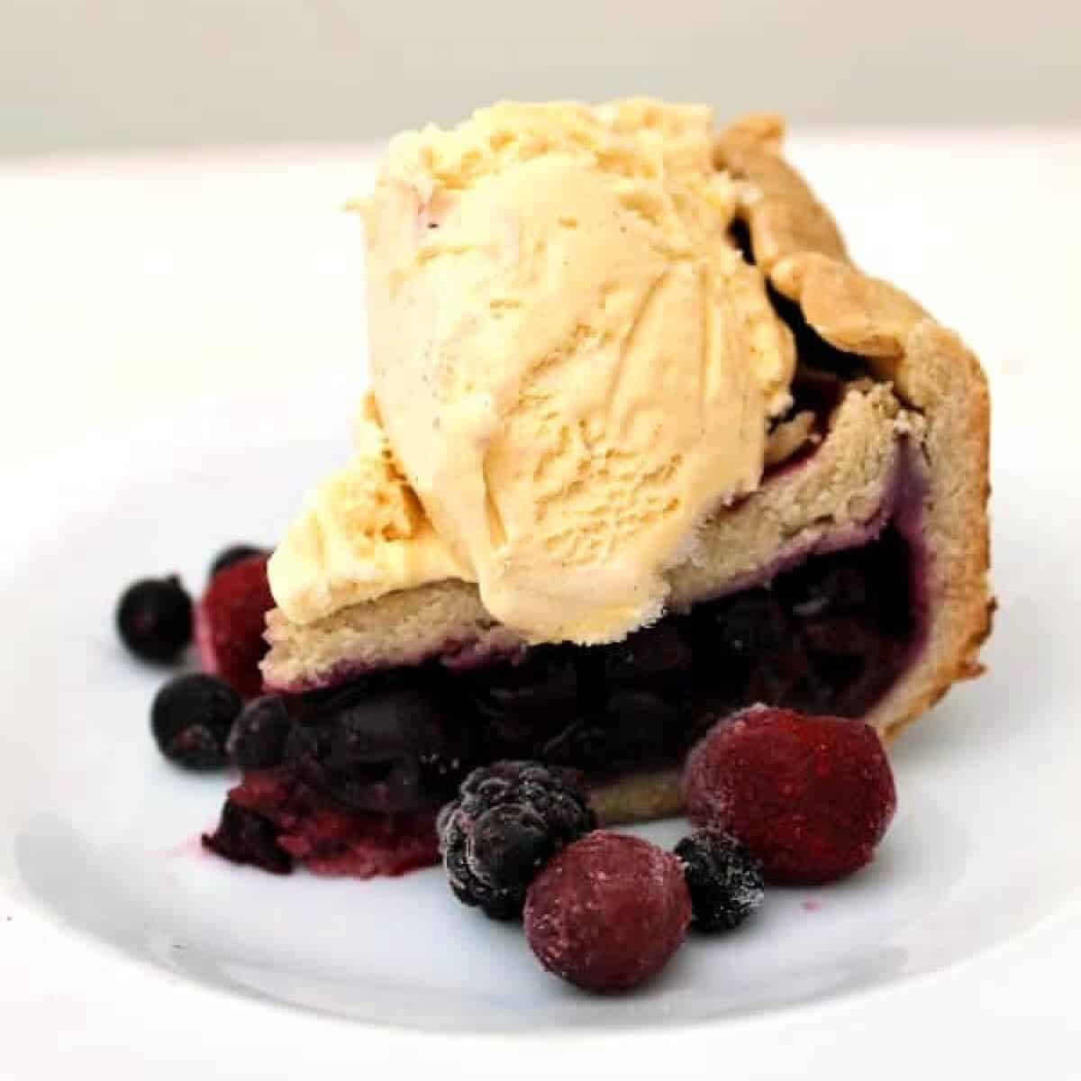 Slice of berry pie topped with ice cream on white plate.
