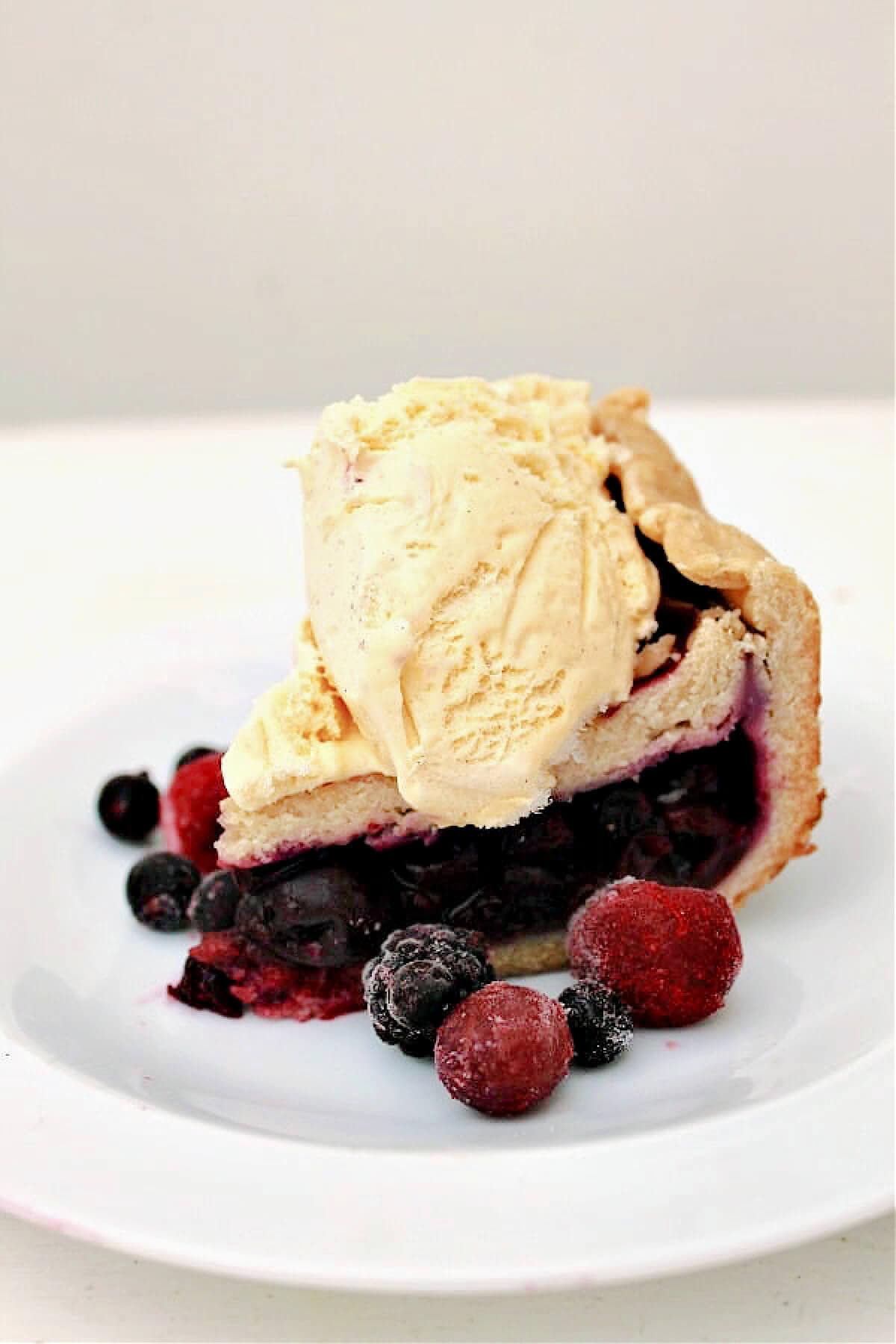 A slice of berry pie topped with a scoop of vanilla ice cream served on a white plate.
