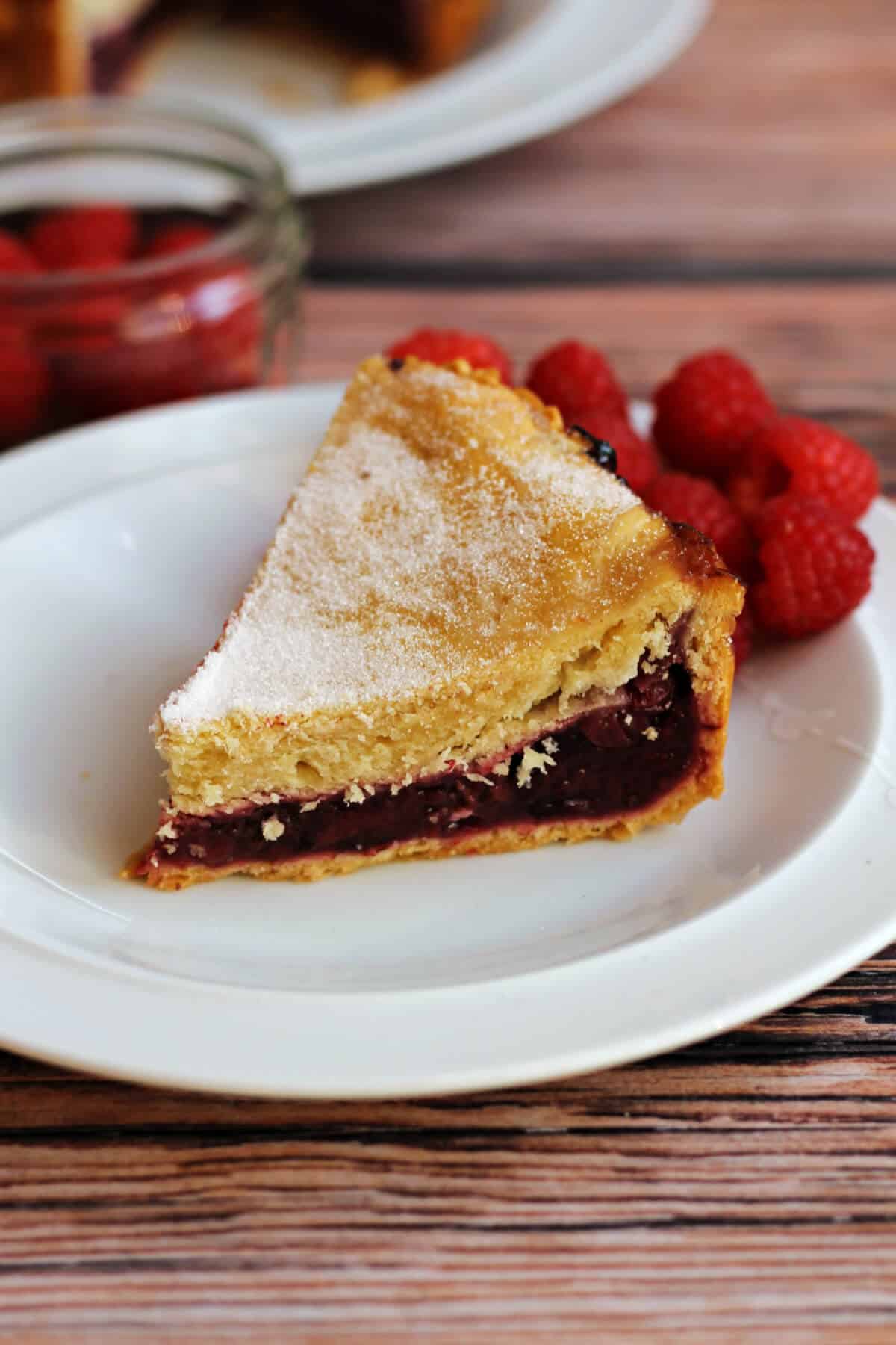 Slice of berry pie dusted with sugar on a white plate, with raspberries.