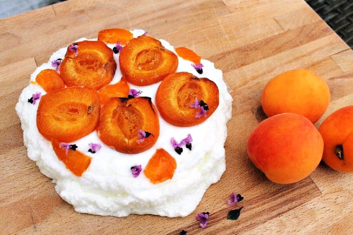 A pavlova decorated with roasted apricots on a wooden board, fresh apricots on the side.