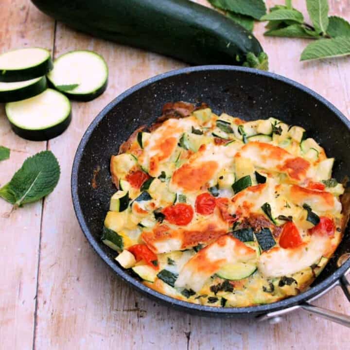 Frittata in a frying pan, with sliced courgettes and herbs around.