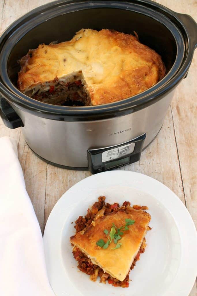 Slow cooker with cooked dish and a plate with a served portion in front.