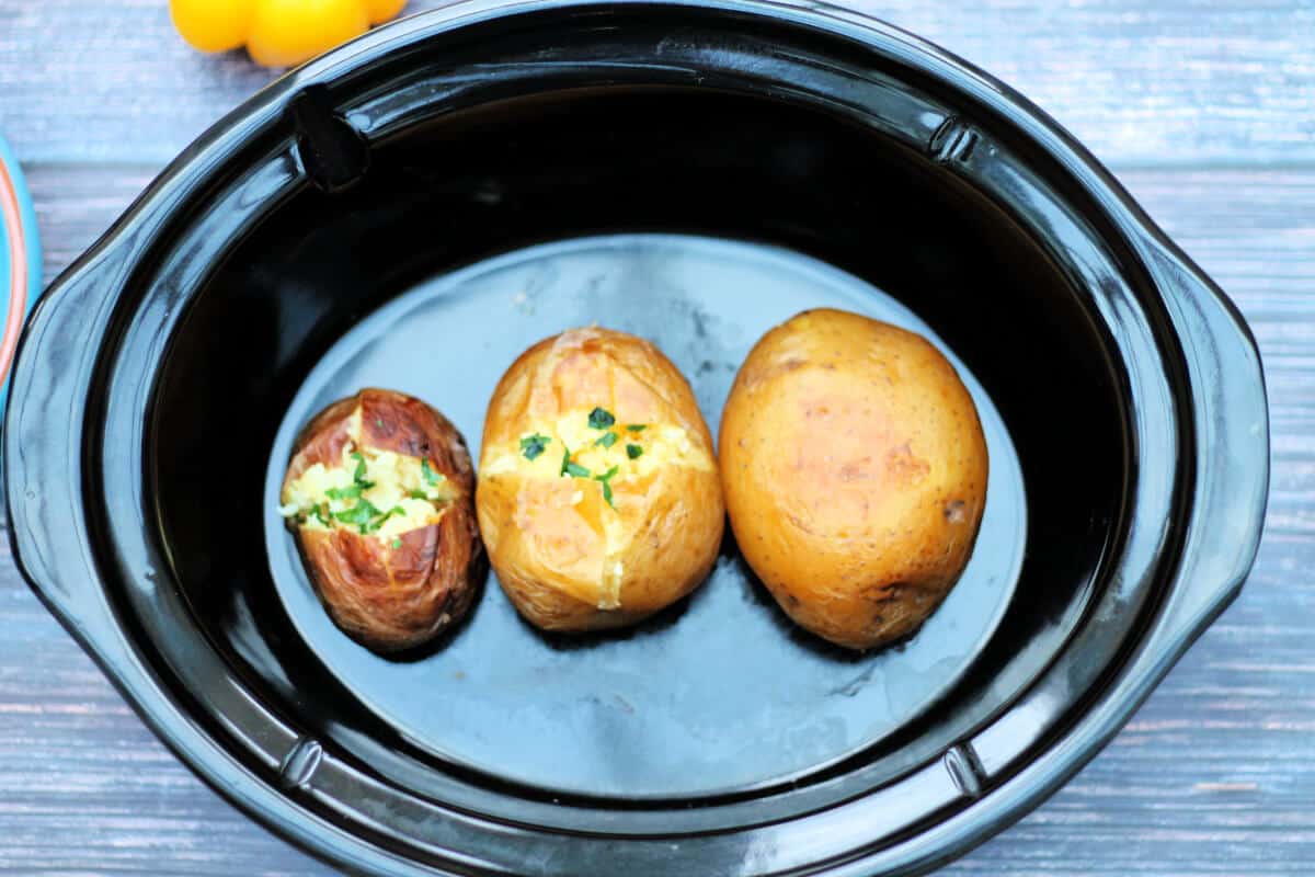 Baked potatoes cut open and topped with herbs in slow cooker pot.