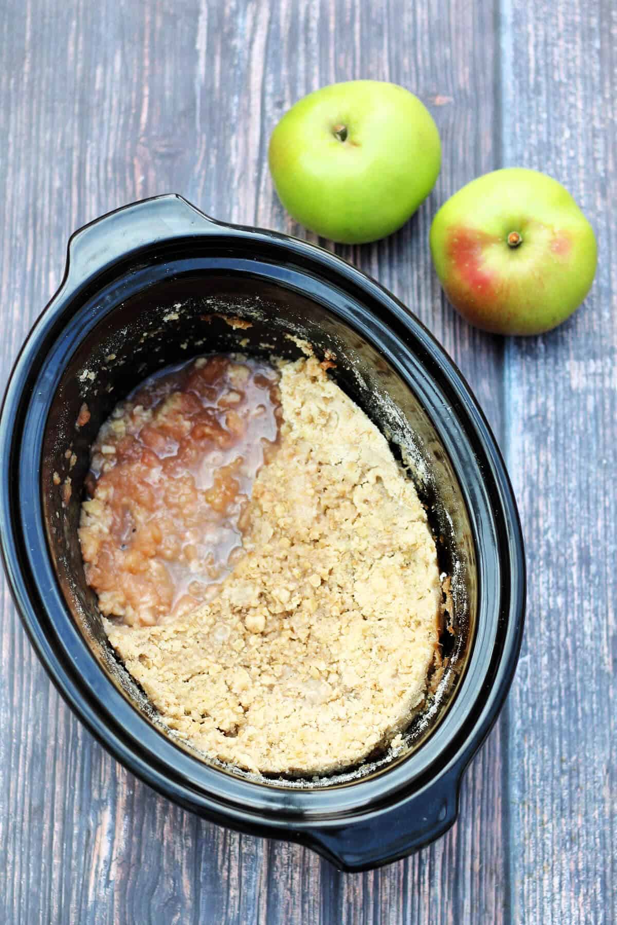 Slow cooker pot with apple crumble, apples behind.