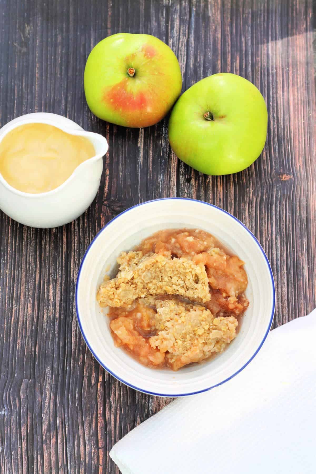 Bowl of crumble with jug of custard and cooking apples.