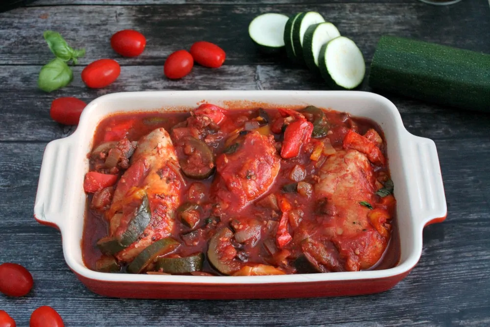 Side view of red serving dish with chicken and tomato stew, with chopped courgette and tomatoes in background.