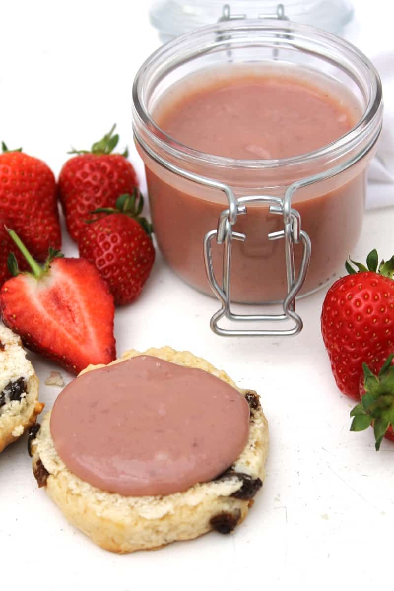 Jar of strawberry curd in background, with half a scone with strawberry curd spread on it in front.