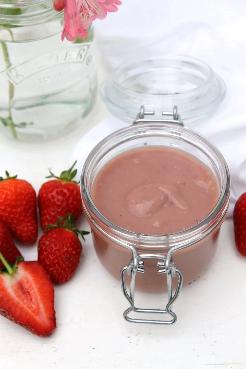 Jar of strawberry curd with strawberries next to it and a small jug of pink flowers in background.