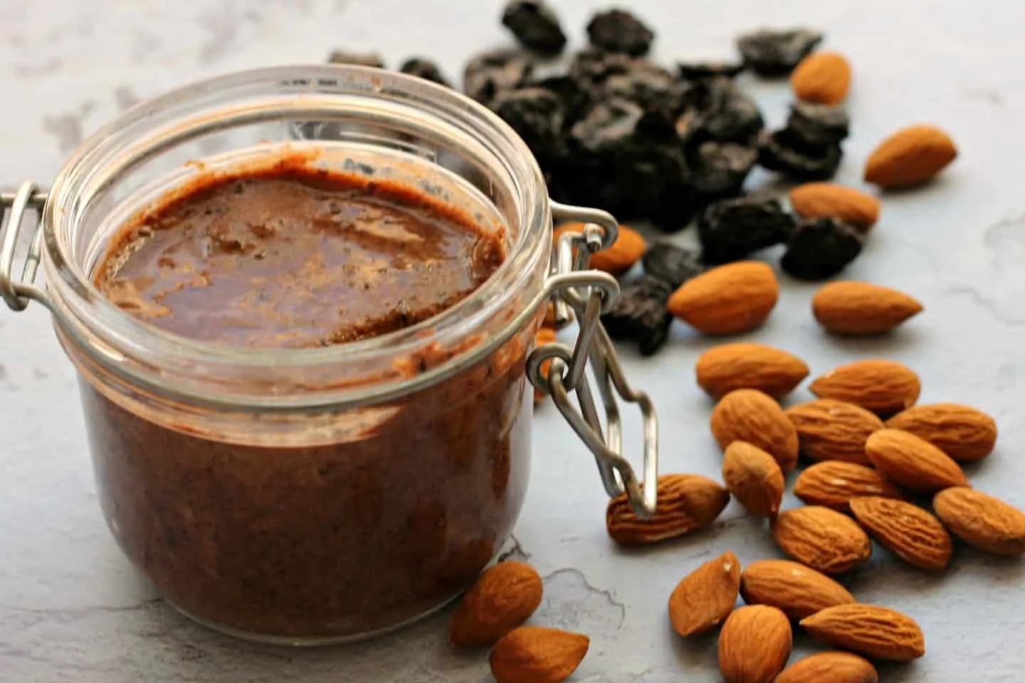 A pot of almond butter on a grey background with almonds and cherries around it.
