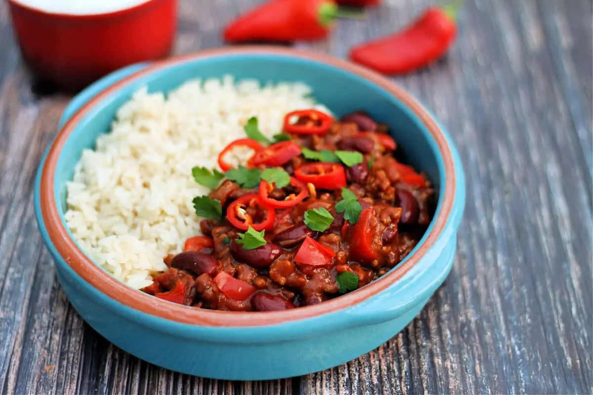 Close up of light blue bowl with chilli and rice, with red chillis in background.