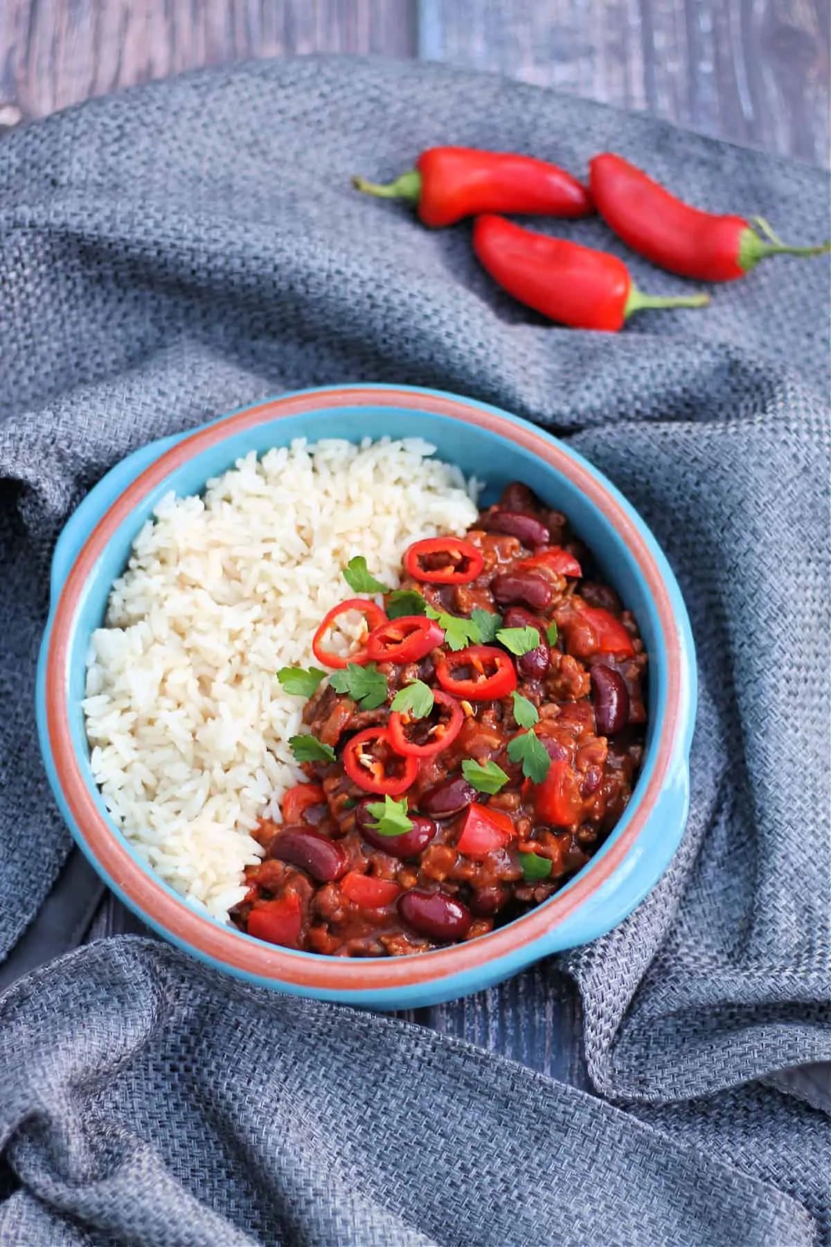 Bowl of chilli and rice with red chillis and herbs on top, on grey fabric background.