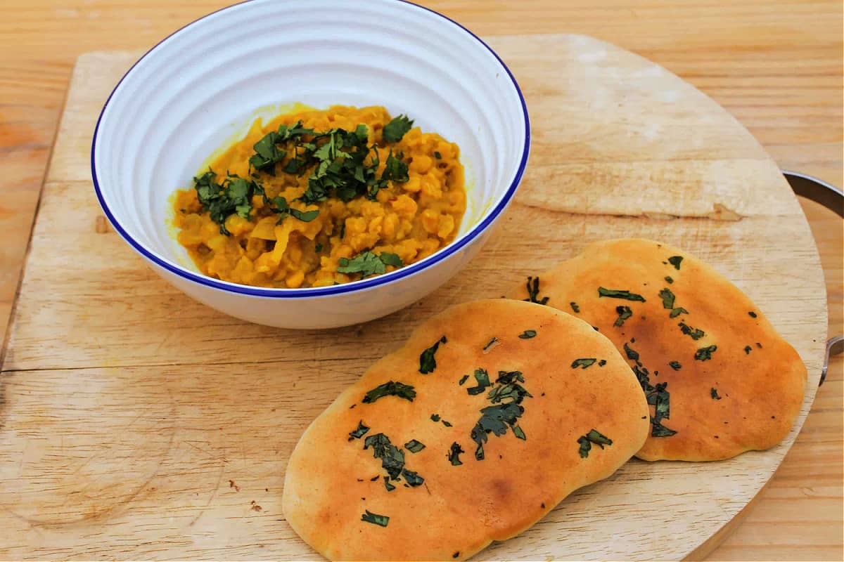 White bowl of yellow dal topped with coriander on a wooden board with homemade naan breads to the side.