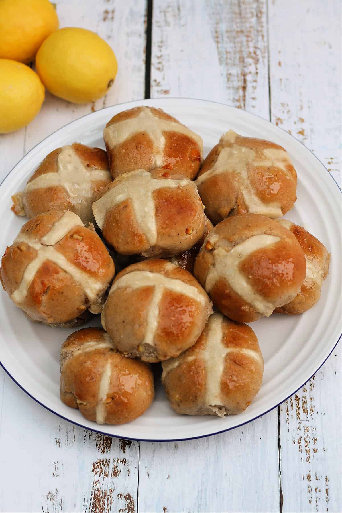 Hot cross buns on a white plate, with lemons in the background. on white wooden surface.