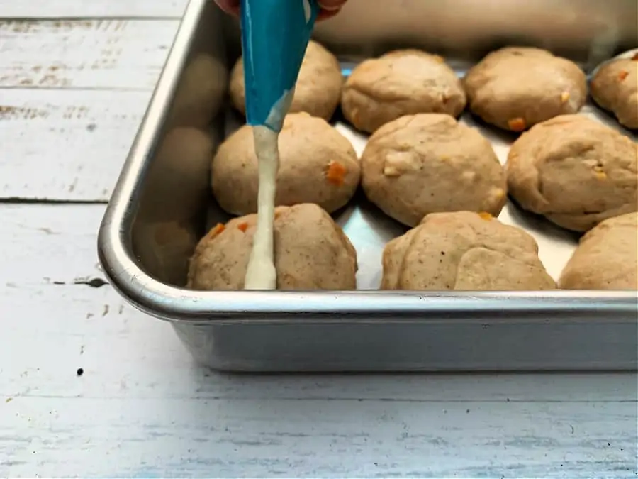 Piping crosses on hot cross buns in a baking pan using a piping bag.