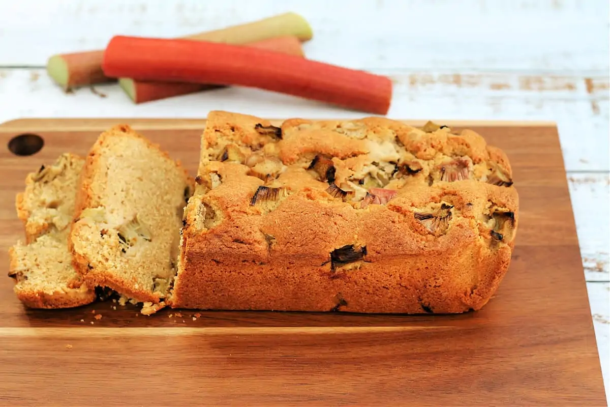 A loaf cake on a wooden board with pieces of rhubarb in the background.