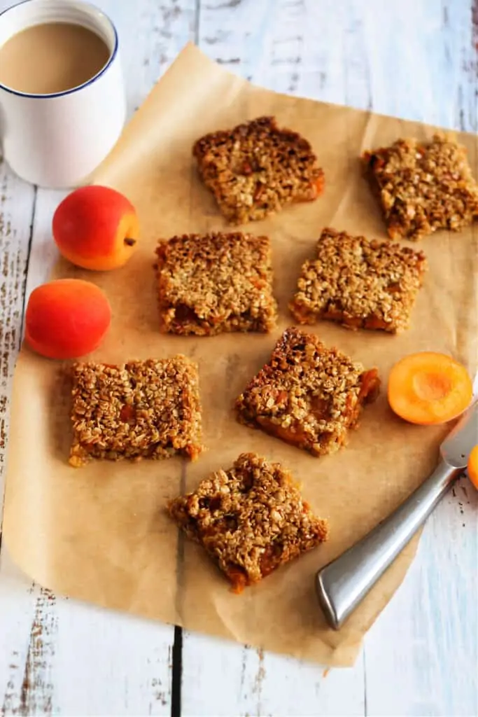 Flapjack pieces on baking paper, with knife, apricots and white cup of tea.