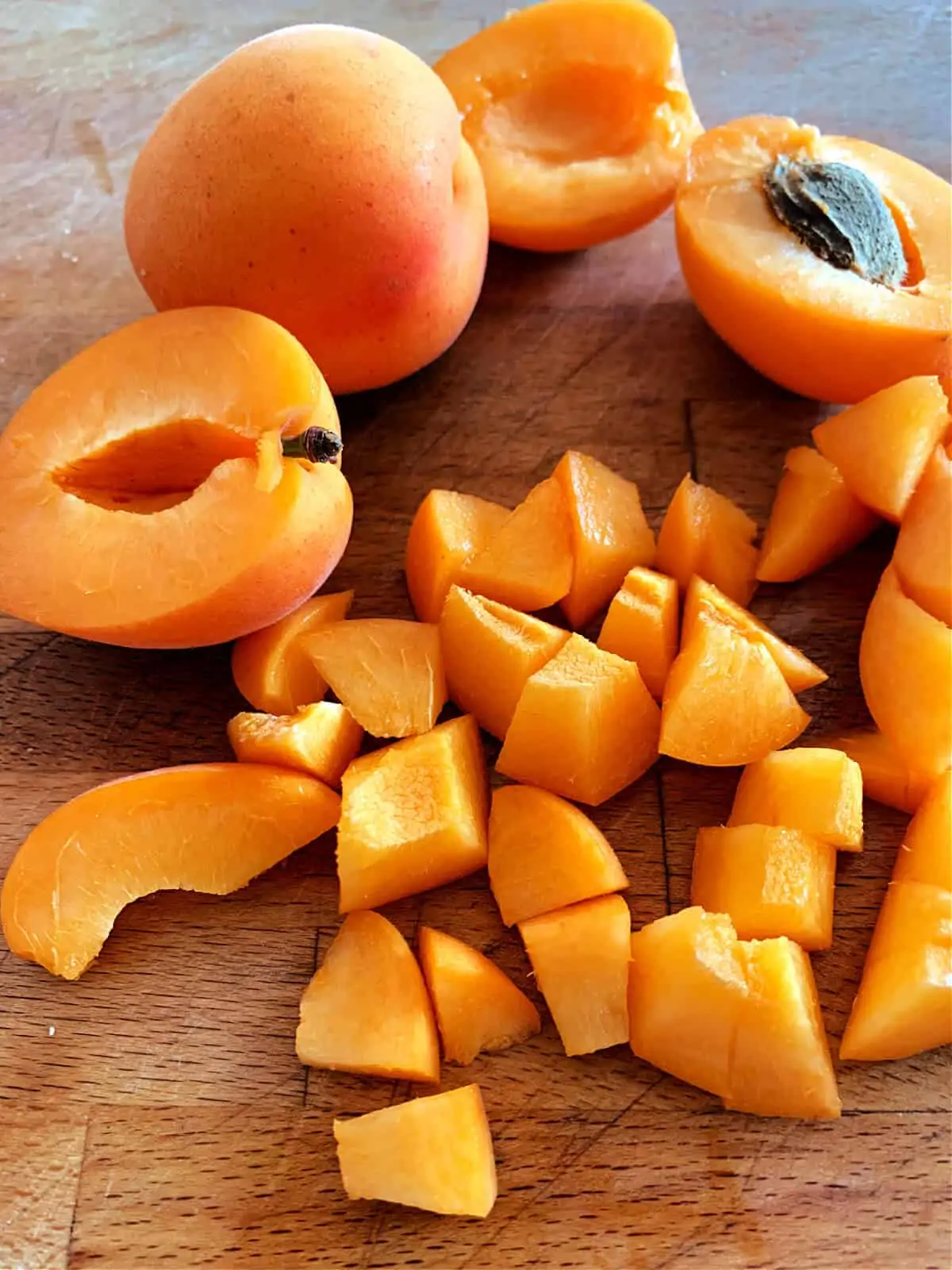 Fresh apricots on a chopping board, some chopped into small pieces, some halved, one whole.
