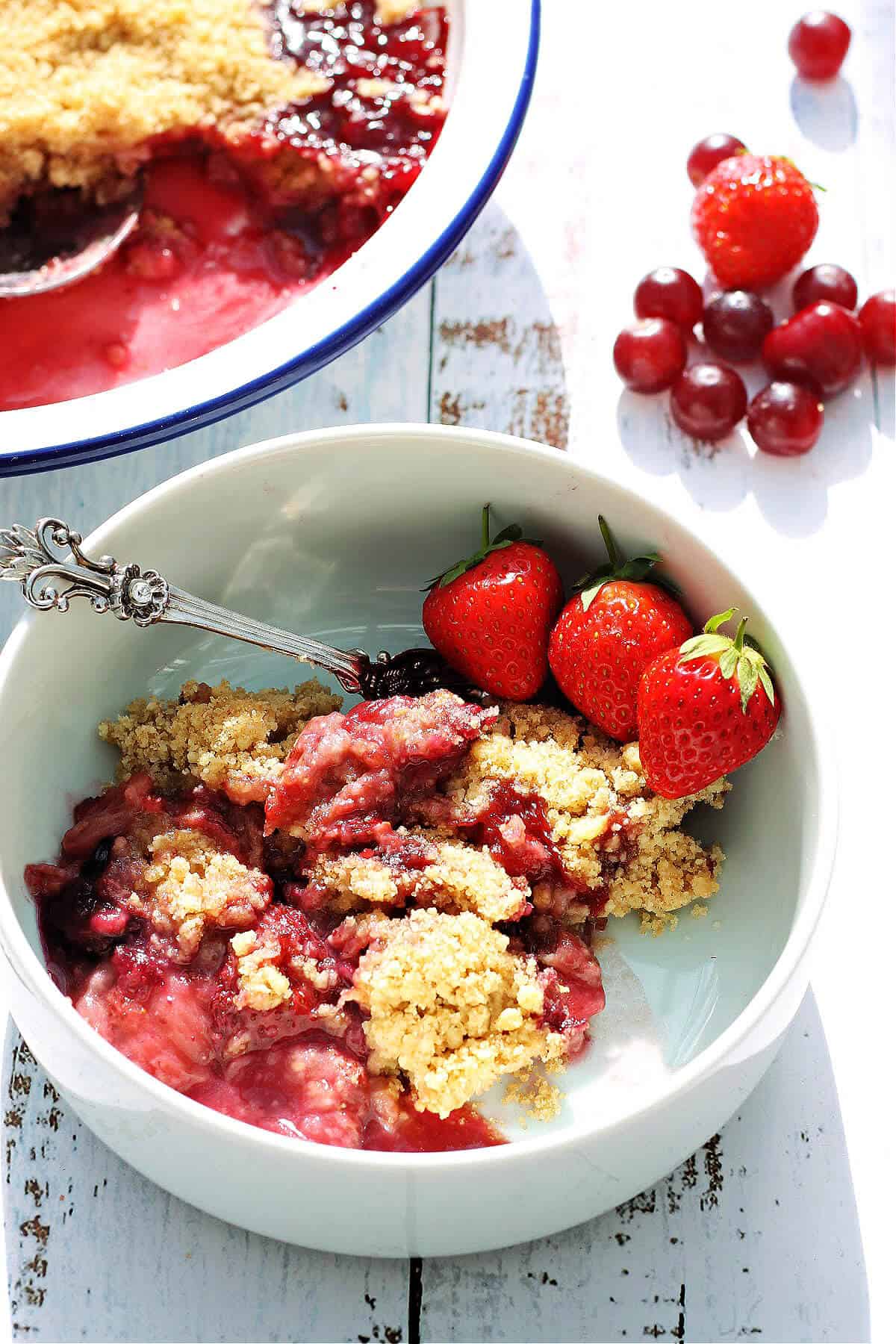 A white bowl of berry crumble with fresh strawberries in the front, serving dish and fresh berries in the background.