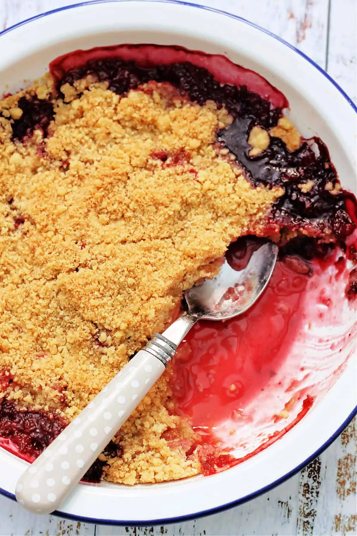 A white ceramic bowl with blue edge containing berry crumble with juices oozing out, with a serving spoon.