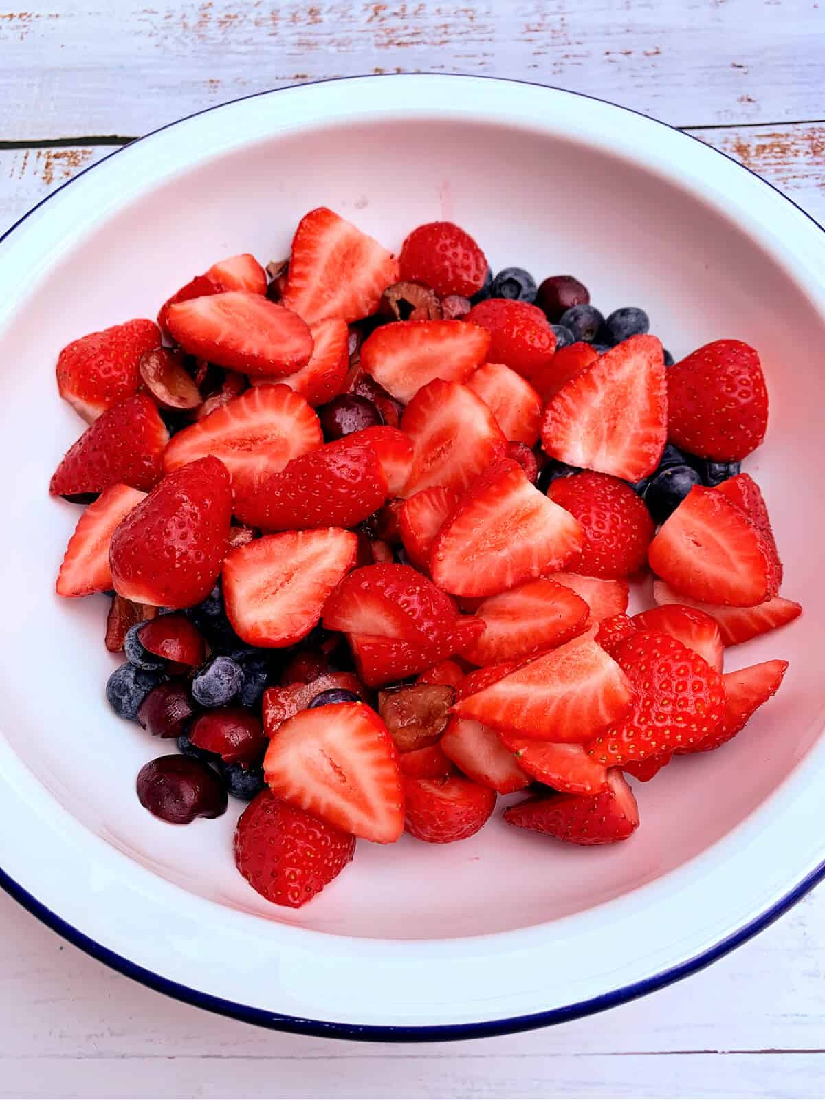 Round white enamel bowl filled with sliced strawberries, cherries and blueberries.