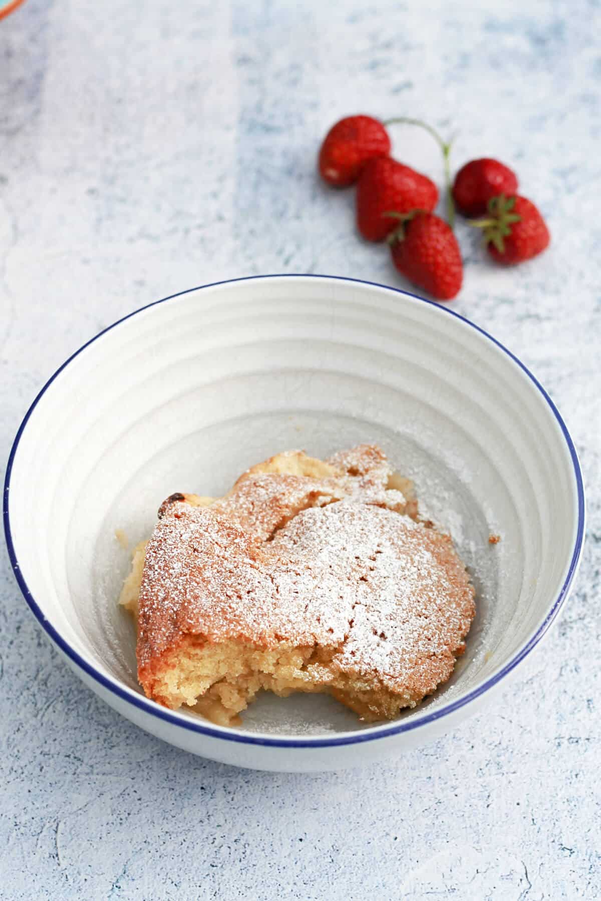 A white bowl with apple sponge dusted with icing sugar, a few strawberries in the background.