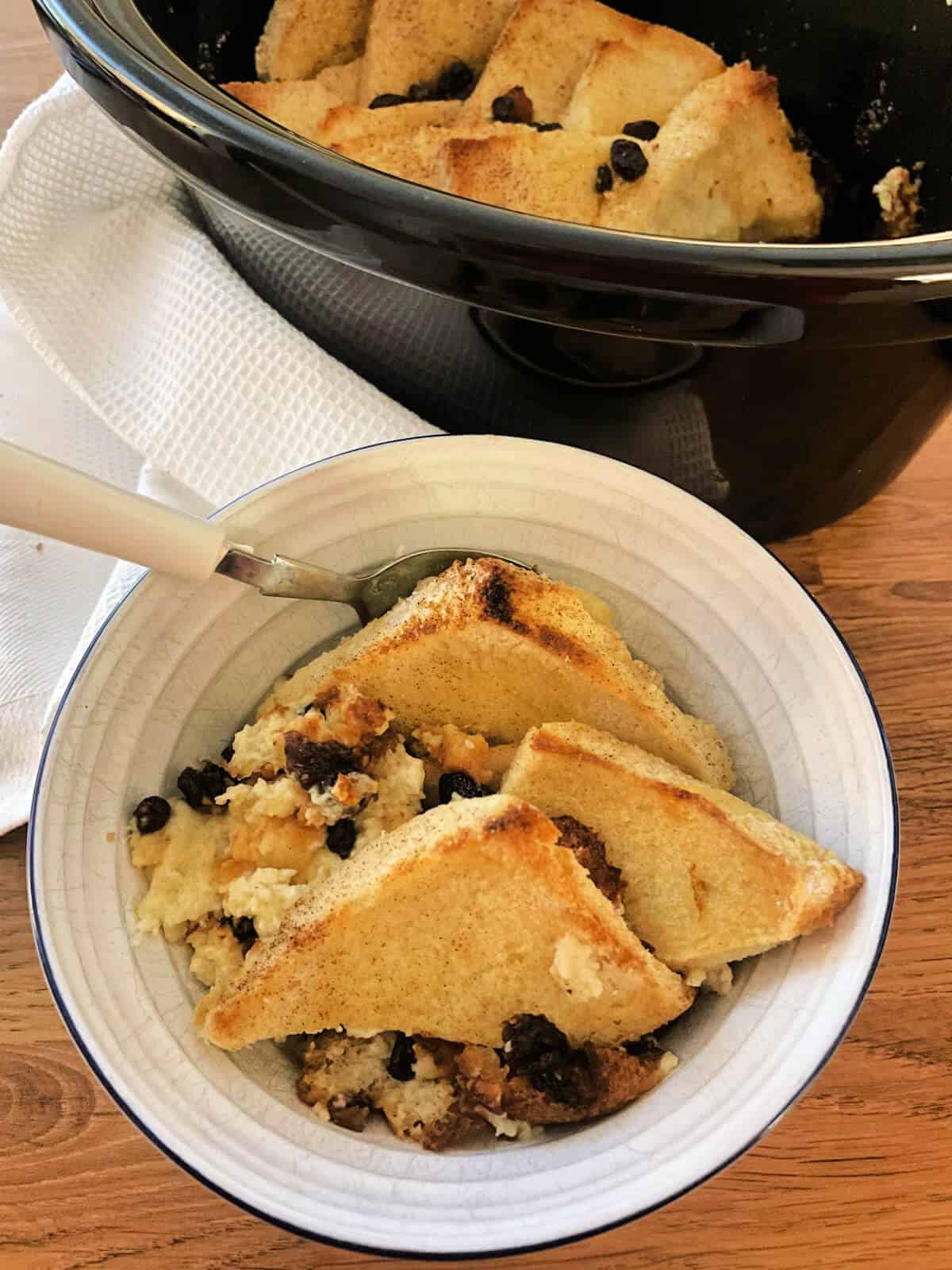 Bowl of bread and butter pudding next to slow cooker serving pot.