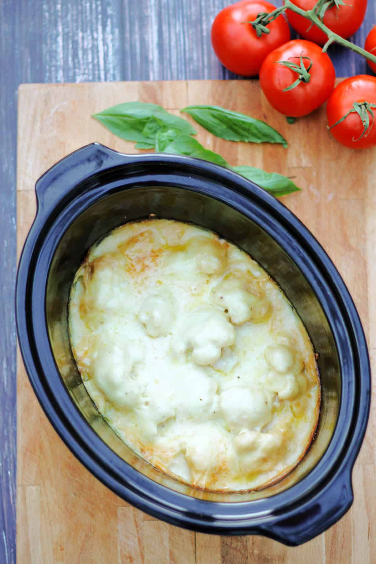 Slow cooker pot filled with cauliflower cheese, tomatoes and basil on side.