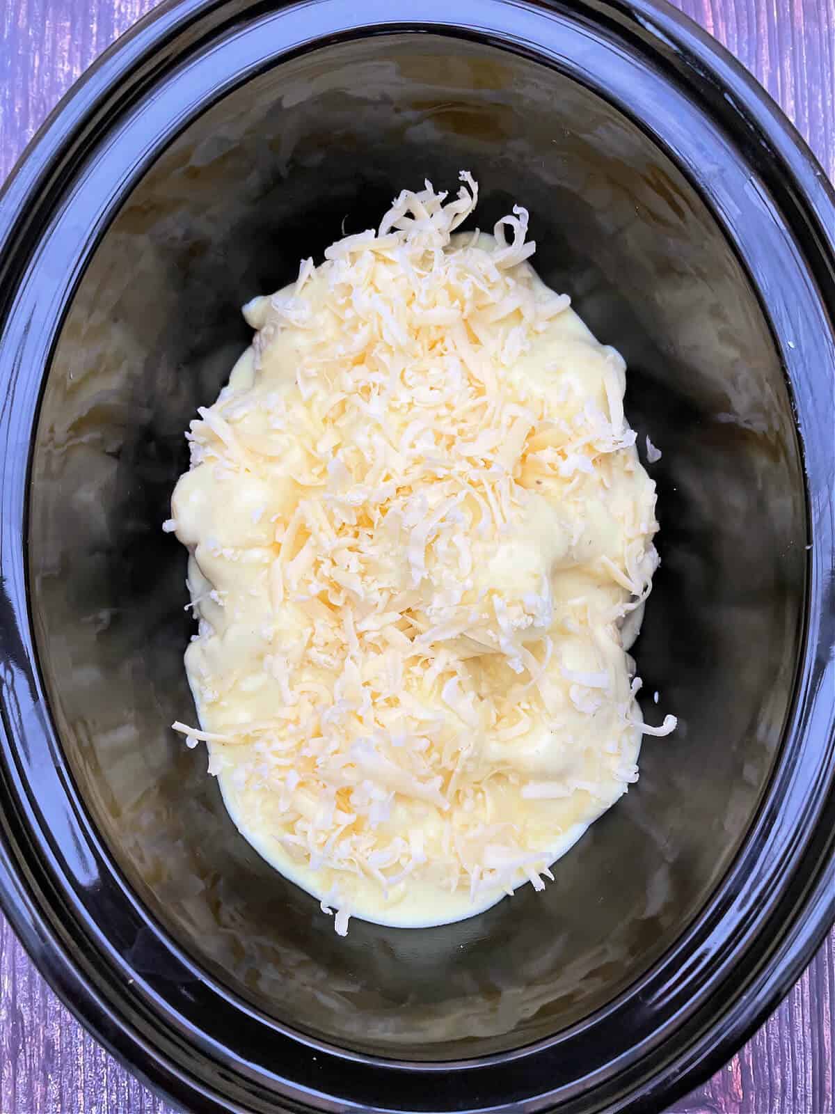Cauliflower cheese mixture with grated cheese on top, in slow cooker pot.