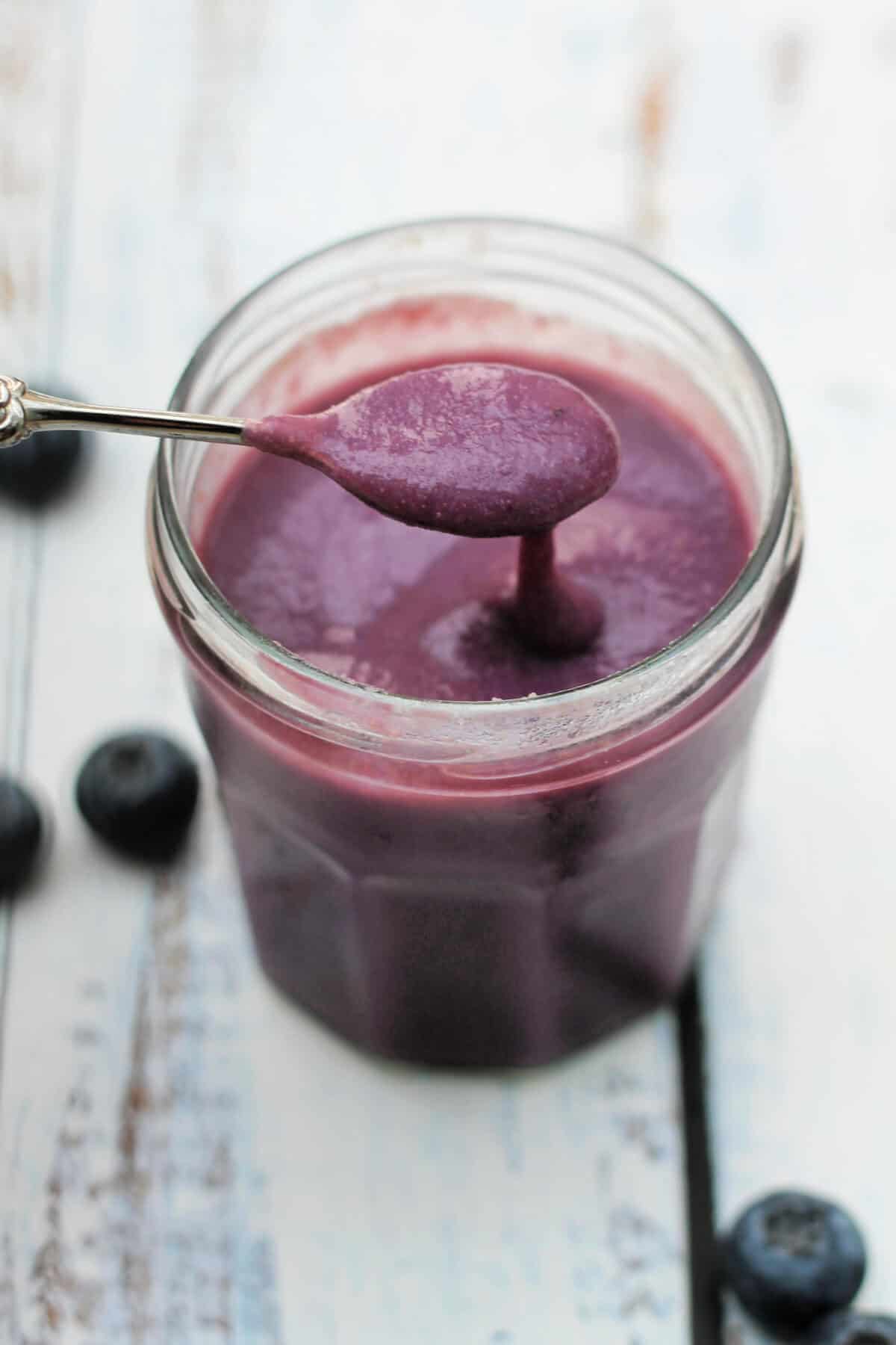 A jar of blueberry curd with a teaspoon above spooning out curd.