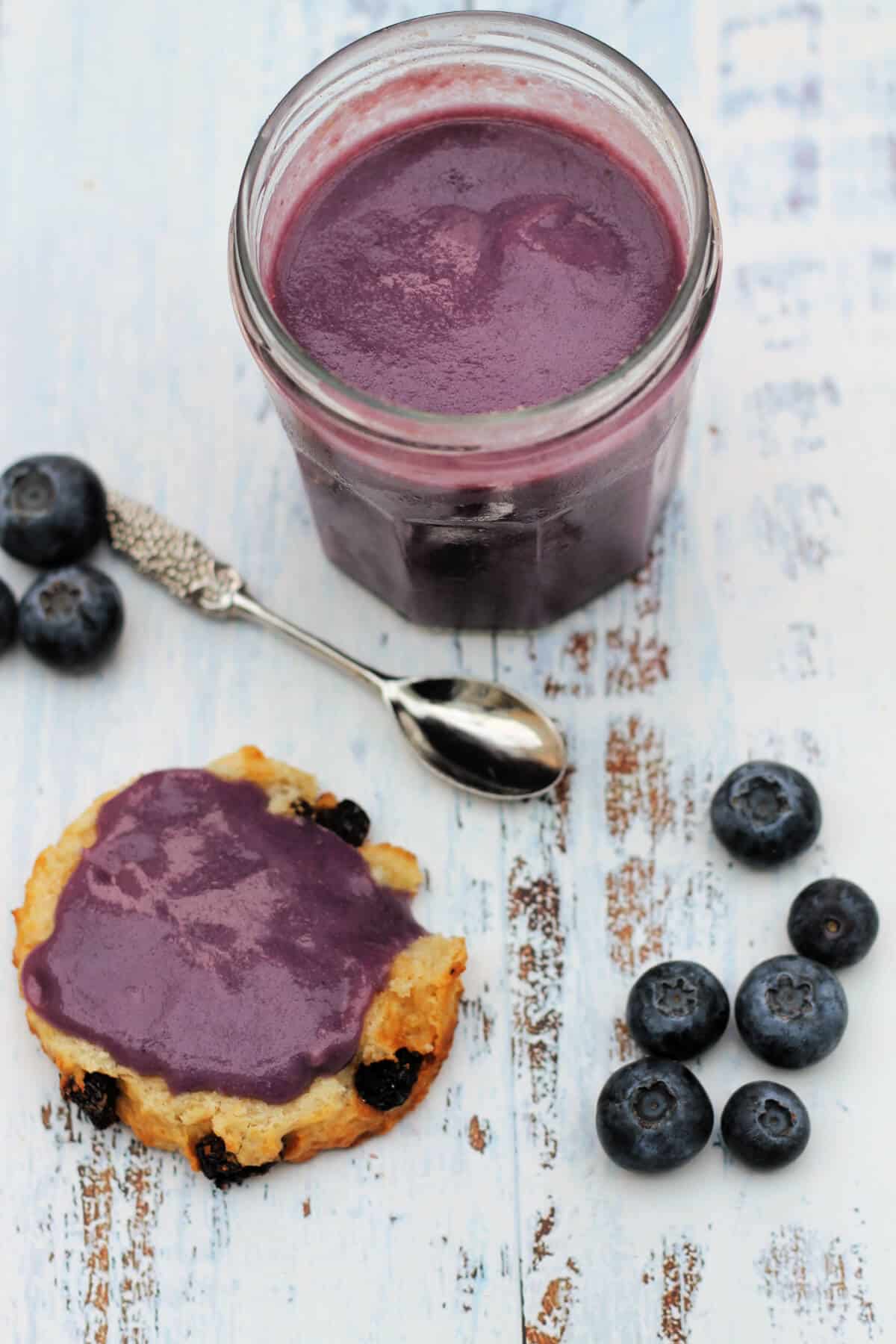 Jar of blueberry curd with a scone spread with curd, ornate spoon and blueberries next to it.