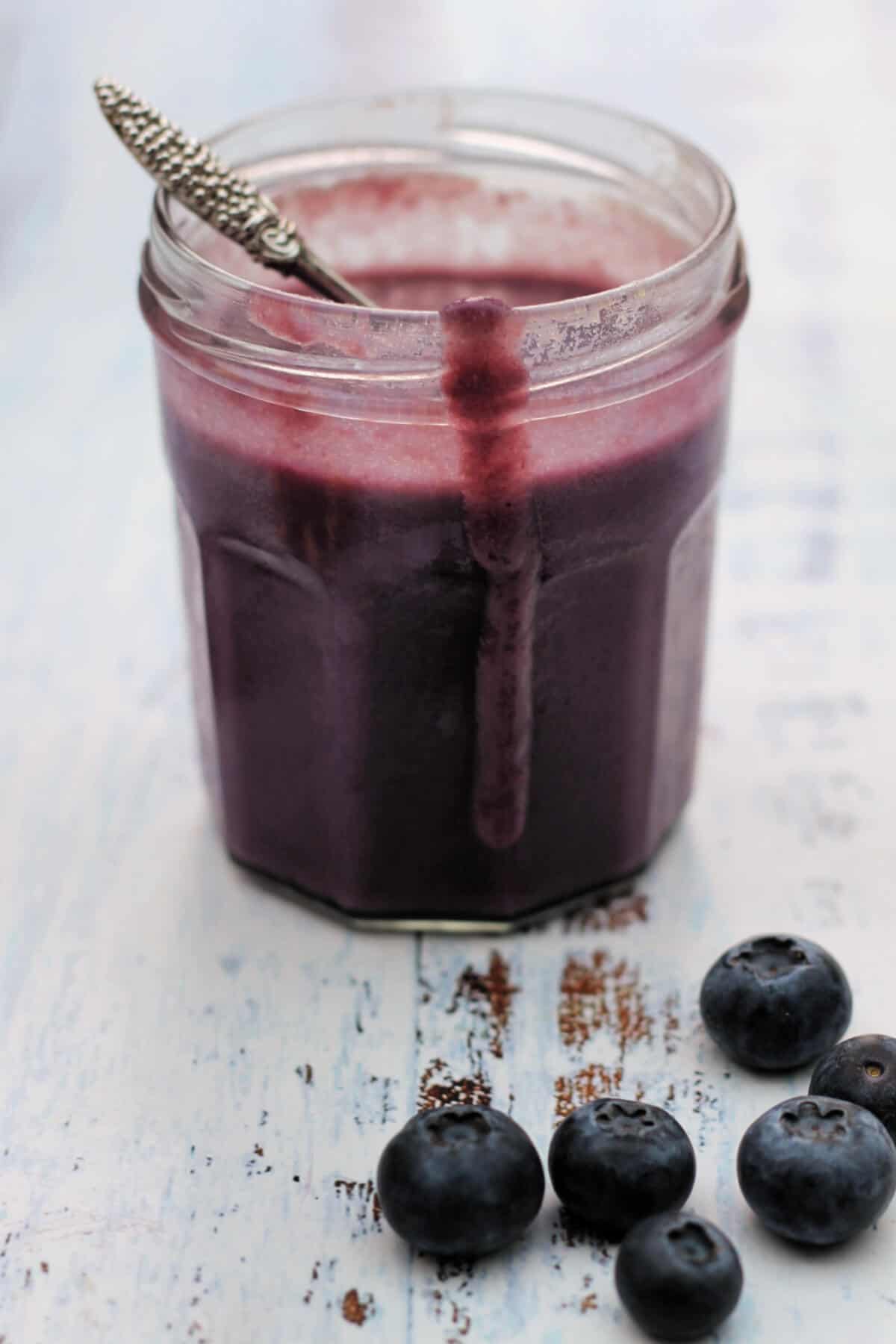 Jar of blueberry curd with drip down the side, spoon in jar, with a few blueberries beside it.