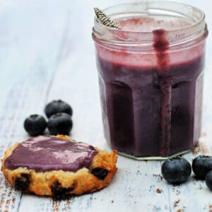 Jar of curd with a drip down the side of it, half a scone spread with blueberry curd beside it.