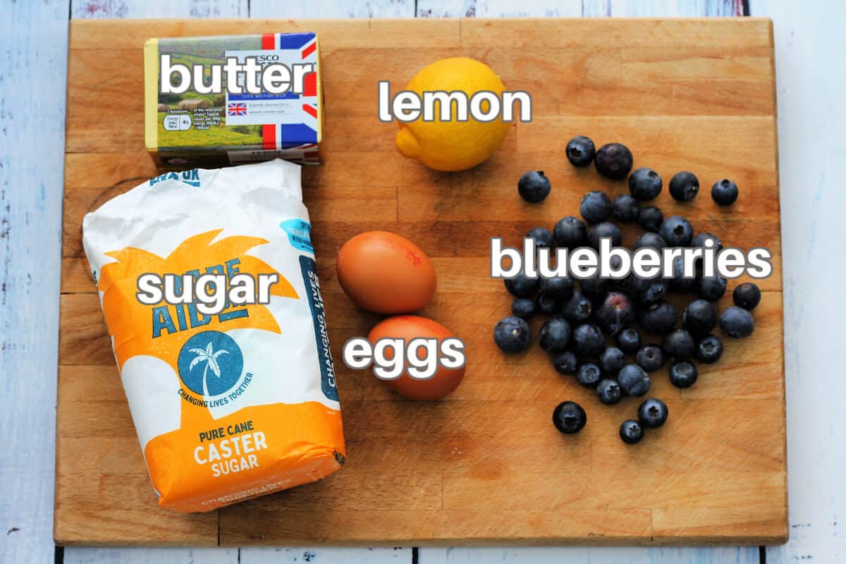 Labelled ingredients on a wooden board - butter, lemon, sugar, eggs and blueberries.