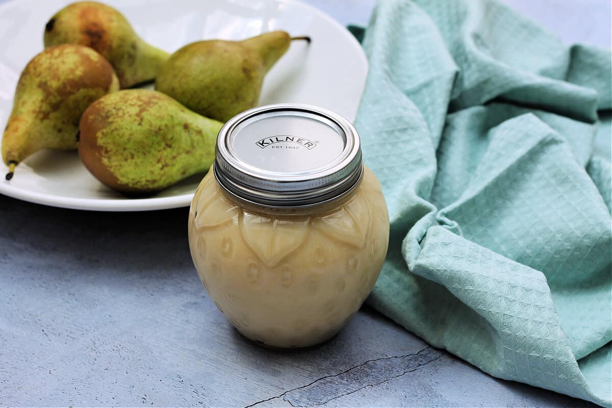 Jar of fruit spread with bowl of pears and green cloth in background.
