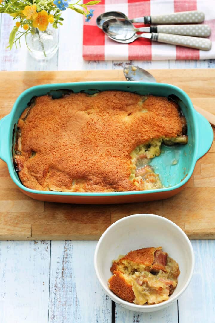 Serving dish with baked rhubarb sponge pudding on a table, bowl of sponge pudding in the front and flowers in a small vase behind.