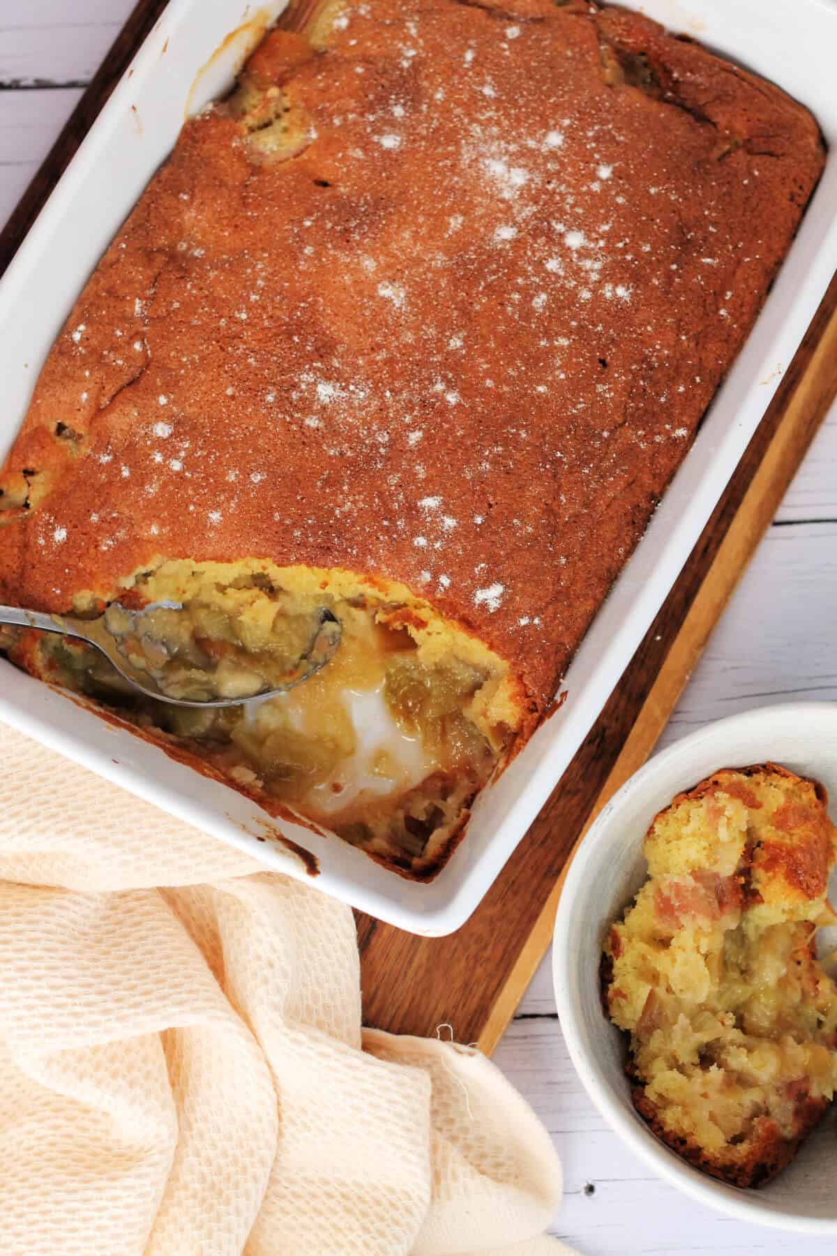 Overhead view of baked sponge with rhubarb layer in white serving dish, with cream tea towel.