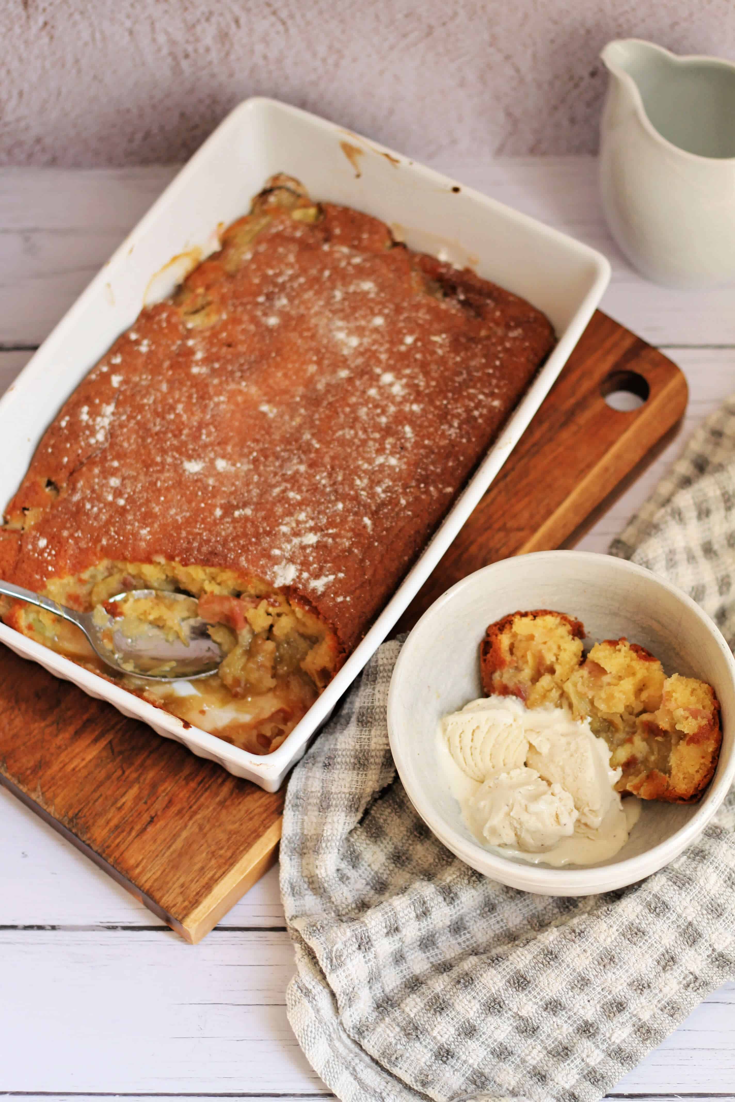 Bowl of rhubarb sponge pudding served with ice cream next to white serving dish, with tea towel.