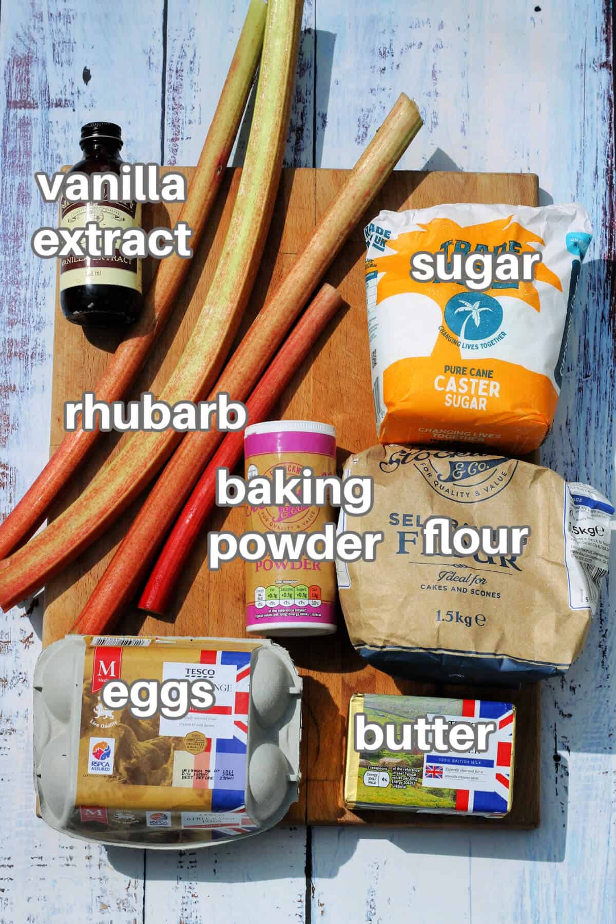Labelled ingredients on a wooden board - rhubarb, sugar, flour, baking powder, eggs, butter, vanilla extract.